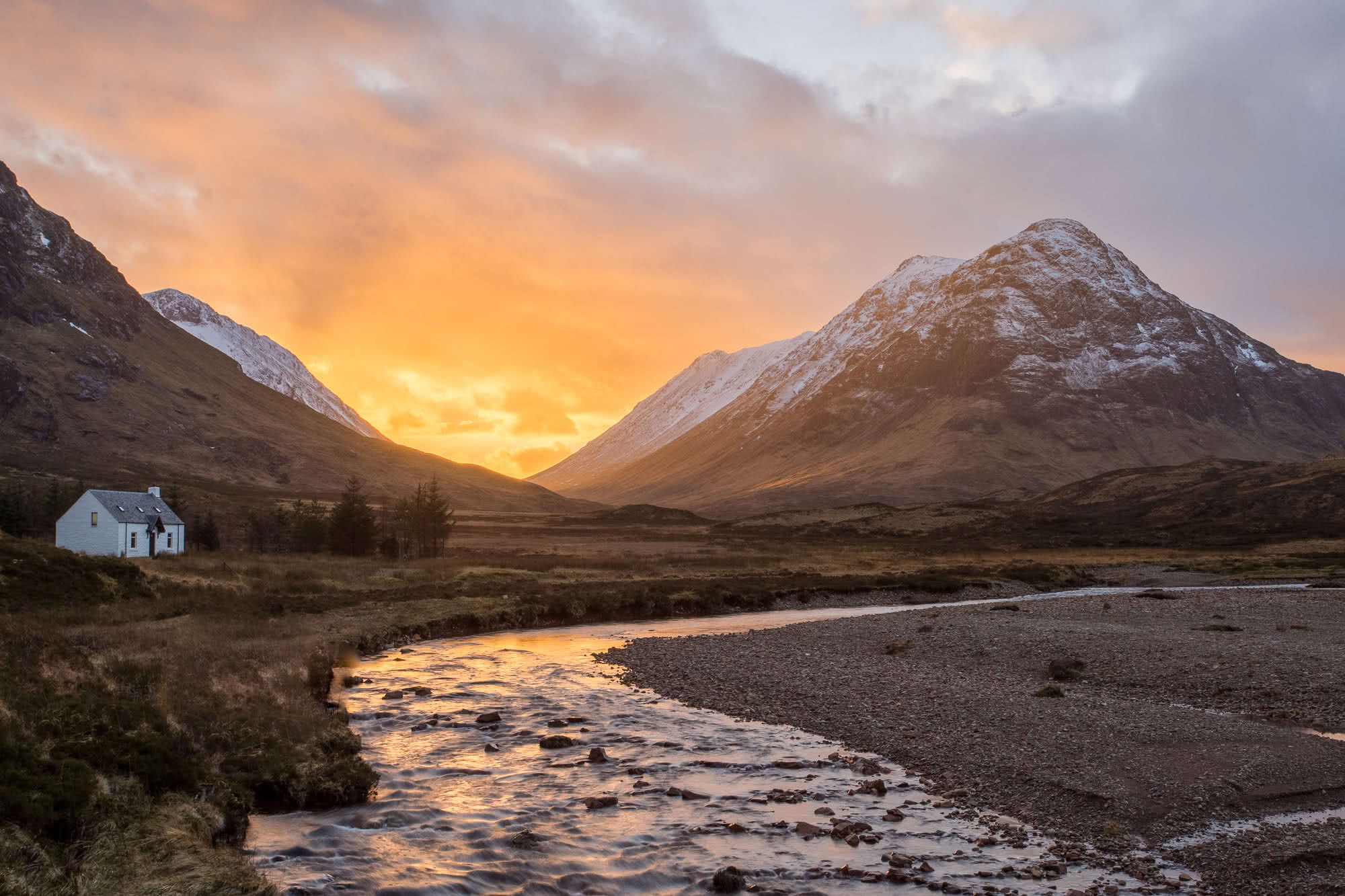 Sunset over Glencoe, Scotland, with a small white cottage beside a winding stream and snow-capped peaks